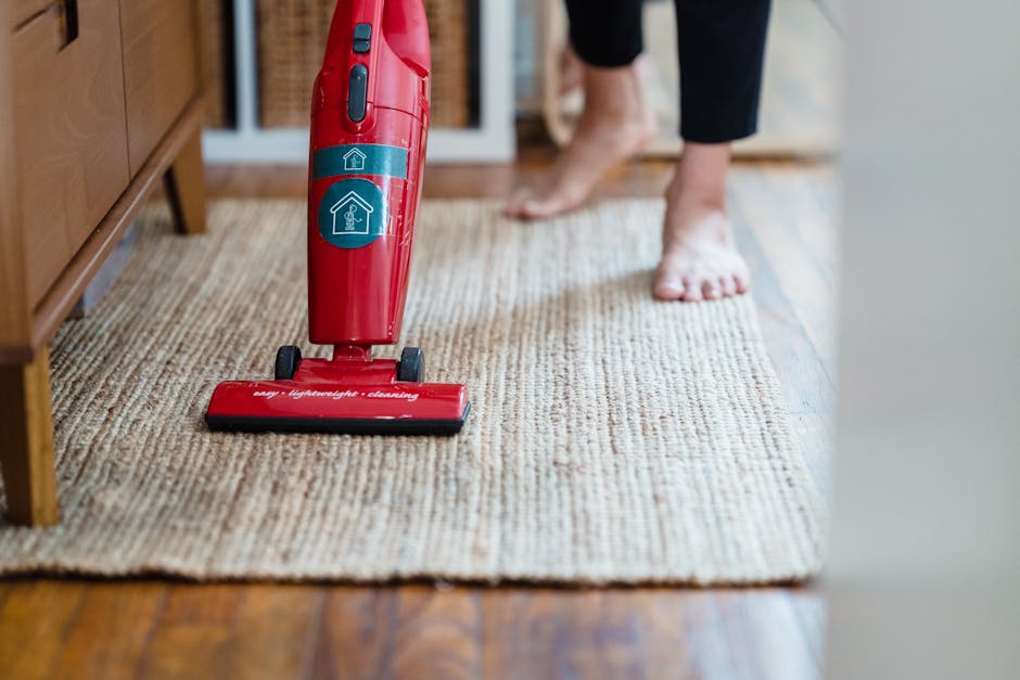 A person vacuuming a beige textured carpet in a residential room, with their feet visible and wearing black pants. The vacuum cleaner is bright red with a digital display and labels indicating 'advanced light-weight cleaning,' positioned in the foreground on the carpet. The room features hardwood flooring along the edges of the carpet and wooden furniture, with soft natural light illuminating the space. The scene exemplifies surface cleaning and maintenance within a home environment, aligning with professional domestic cleaning standards promoted by GreenwichCarpetCleaning.co.uk.