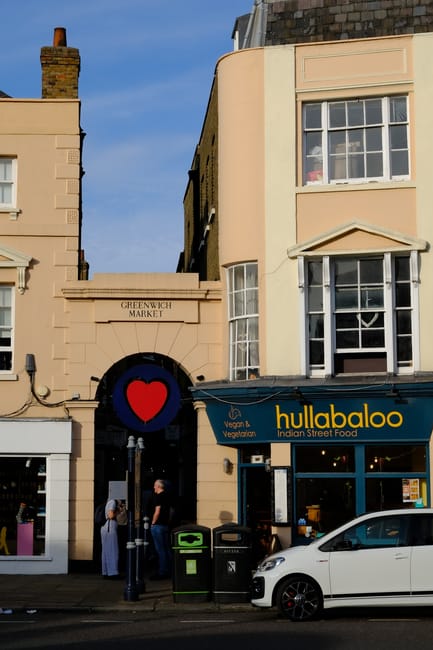 Exterior view of a building in Greenwich Market area featuring a residential window with multiple panes, a sign for Hullabaloo Indian Street Food restaurant, and a decorative entrance archway with a red heart logo above the doorway. In front, there is a white car parked on the street, along with a green recycling bin and trash bin. The building's facade is light beige with contrasting darker upper sections, and the scene is illuminated by natural daylight, showcasing a clean and well-maintained urban environment.