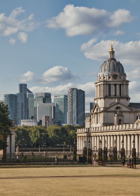 A wide-angle view of a historic domed building with classical architecture, featuring tall columns and ornate details, situated in a park with a low fence and benches in the foreground. Behind it, modern skyscrapers with glass facades reflecting the sky create a contrasting cityscape. The area is illuminated by natural daylight, with a partly cloudy sky overhead. The scene captures a blend of historic and contemporary architecture, emphasizing the clean and well-maintained appearance of the surroundings, aligning with the concepts of surface cleaning and sanitisation relevant to the Best end of tenancy cleaning in Greenwich Market area, GREENWICH. The image is associated with GreenwichCarpetCleaning.co.uk's cleaning services.