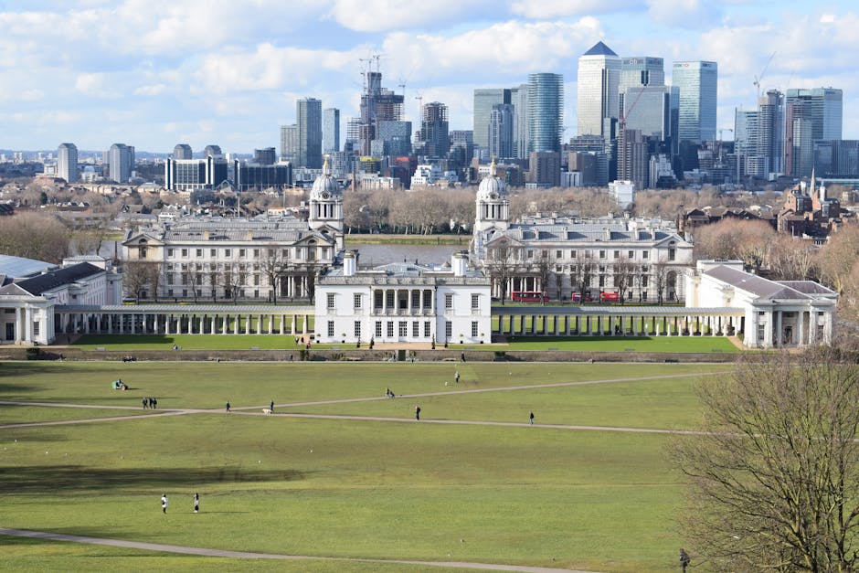 A panoramic view of a historic white palace with classical architecture, featuring columns and domed towers, situated in a large landscaped park with well-maintained grassy areas and walking paths. The palace is set against a backdrop of modern high-rise office buildings and skyscrapers in the city skyline of Greenwich. The scene is illuminated by natural daylight, and the park appears clean and orderly, with a few people strolling or relaxing on the grass, reflecting maintenance and tidiness. The image may include visible tools or signs of recent cleaning activities, emphasizing thorough surface cleaning, deep cleaning, and sanitisation by Greenwich Carpet Cleaning, highlighting its domestic cleaning services in the Greenwich area.