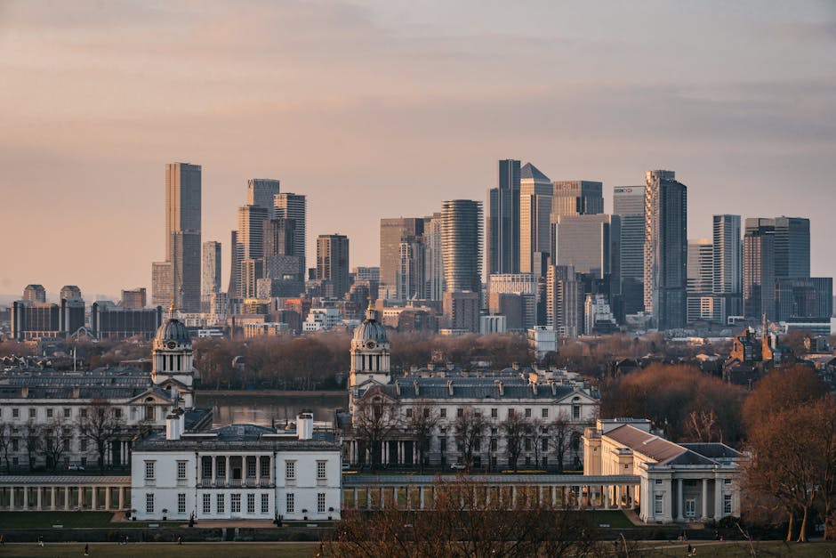 A panoramic view of the Greenwich skyline featuring modern glass skyscrapers and high-rise office buildings in the background, illuminated by soft natural light during sunset. In the foreground, there is a historic white building with classical architecture, topped with domed clock towers, surrounded by leafless trees and well-maintained grassy areas. The scene captures the contrast between contemporary urban development and historic architecture, with the cityscape appearing clean and well-preserved, reflecting the importance of surface cleaning and maintenance services provided by Greenwich Carpet Cleaning in the Greenwich area.