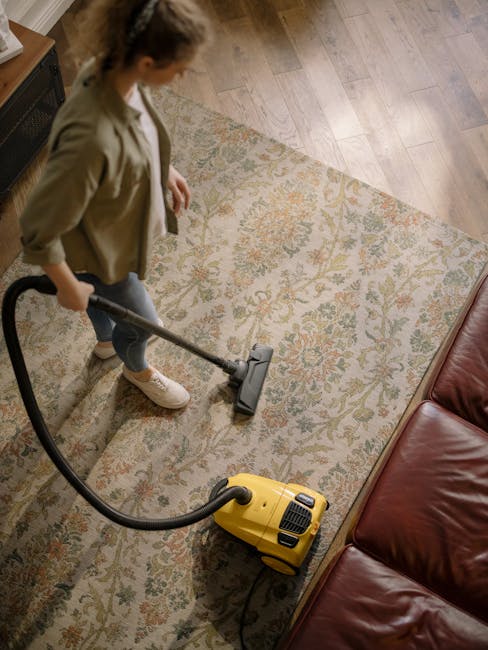 A woman vacuuming a patterned area rug in a living room with hardwood flooring and a leather sofa. The rug features intricate floral designs in muted tones of beige, green, and orange. She is using a yellow and black canister vacuum cleaner, holding the hose and cleaning the surface thoroughly. The room is brightly lit by natural light, highlighting the clean and well-maintained appearance of the space. This scene illustrates surface cleaning and deep cleaning practices typical of domestic cleaning services, as promoted by GreenwichCarpetCleaning.co.uk, to maintain hygienic and tidy SE10 homes in Greenwich.