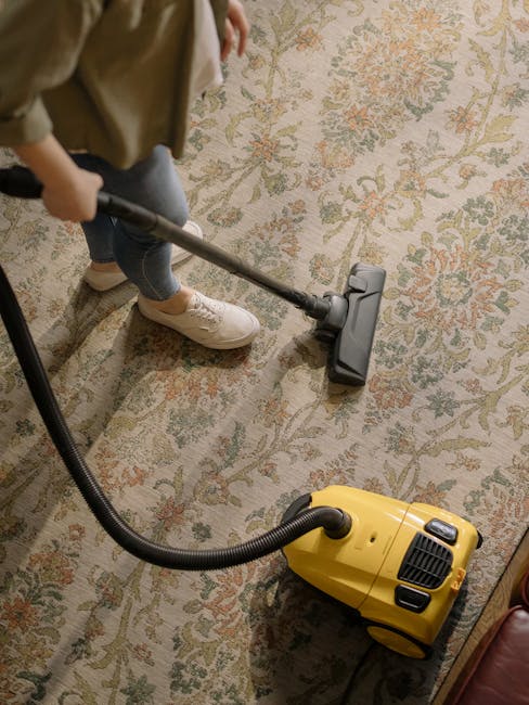 A person using a handheld vacuum cleaner connected to a yellow canister vacuum to perform surface cleaning on a patterned, beige and green rug in a domestic setting. The individual, wearing casual clothing and beige shoes, is guiding the vacuum over the carpet that features floral motifs. The scene illuminates the room with soft natural light, highlighting the cleanliness and maintained condition of the rug. This image demonstrates thorough deep cleaning processes, reflecting hygienic standards in residential carpet maintenance, consistent with service offerings by GreenwichCarpetCleaning.co.uk.