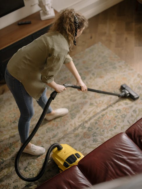 A woman vacuuming a patterned area rug in a living room with hardwood flooring and a leather sofa. The rug features intricate floral designs in muted tones of beige, green, and orange. She is using a yellow and black canister vacuum cleaner, holding the hose and cleaning the surface thoroughly. The room is brightly lit by natural light, highlighting the clean and well-maintained appearance of the space. This scene illustrates surface cleaning and deep cleaning practices typical of domestic cleaning services, as promoted by GreenwichCarpetCleaning.co.uk, to maintain hygienic and tidy SE10 homes in Greenwich.