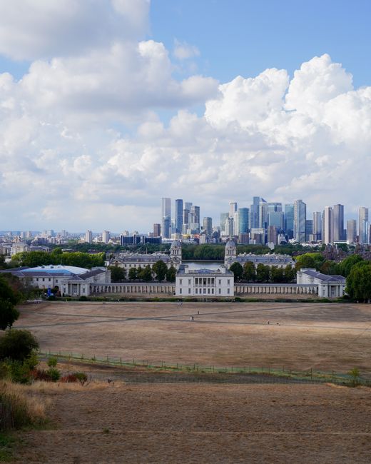 A wide panoramic view of a city skyline with modern glass skyscrapers under a partly cloudy sky, seen from a distance across an open field with dry grass and scattered greenery. In the middle ground, there is a large, white, classical-style building with columns, surrounded by low trees. The scene is bright with natural daylight, showcasing the urban landscape against a backdrop of fluffy white clouds and patches of blue sky. This image emphasizes the contrast between natural open space and the dense, high-rise buildings of the city, illustrating the urban environment from an open, spacious perspective. Greenwich Carpet Cleaning offers expert surface cleaning and deep cleaning services suitable for domestic and commercial spaces, helping maintain hygiene and cleanliness in various environments.