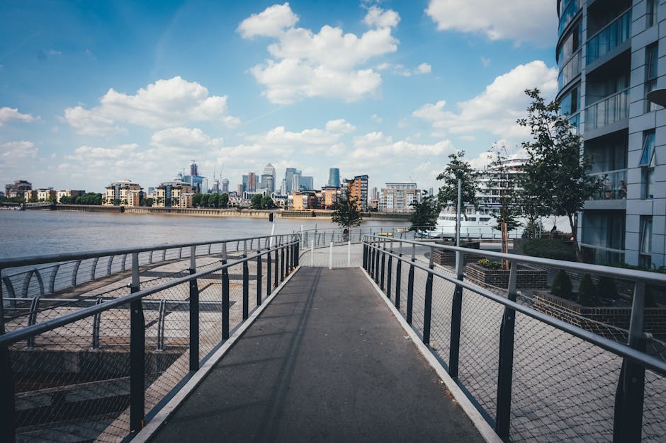 A wide outdoor walkway with metal railing and chain-link fencing along a riverfront, leading towards a city skyline with modern high-rise buildings under a partly cloudy sky. The walkway surface appears clean and well-maintained, with some shadows cast by nearby trees and surrounding structures including contemporary residential buildings with glass balconies on the right side. The scene features natural lighting highlighting the clarity of the water and the urban landscape in Greenwich, aligning with domestic cleaning and surface sanitisation themes, and demonstrates the clean, orderly environment that GreenwichCarpetCleaning.co.uk emphasizes in its deep cleaning services for flats on King William Walk Greenwich.