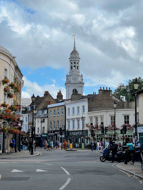 A busy street scene in Greenwich featuring a historic white clock tower with a pointed spire in the background. The street is lined with shops displaying colorful window displays and hanging flower baskets. Pedestrians walk along the sidewalks, and vehicles, including motorcycles and cars, are visible at the intersection. Overhead, the sky is partly cloudy, with patches of blue sky peeking through. The scene is well-lit with natural daylight, capturing a vibrant, lively atmosphere in a historic town center, emphasizing the need for thorough cleaning and maintenance of the urban surfaces by Greenwich Carpet Cleaning for settings like this.