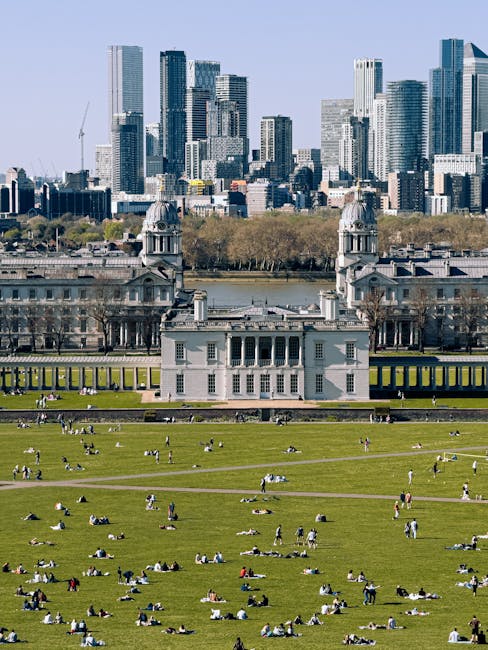 A wide view of Greenwich Park with lush green lawns and numerous people relaxing, sitting, and walking on the grass. In the background, the historic Queen's House with its distinctive white façade and classical architecture is visible, featuring symmetrical windows and two prominent towers on either side. Behind the Queen's House, the River Thames runs through the scene, with a line of trees along the riverbank. The modern city skyline of Greenwich with tall skyscrapers and office buildings rises in the distance under a clear blue sky. The outdoor scene is well-lit, suggesting a sunny day, and the overall impression emphasizes the cleanliness and maintenance of the park’s surfaces, aligning with the themes of deep cleaning and surface sanitisation by Greenwich Carpet Cleaning, as referenced in the page titled 'Same day carpet cleaning near Greenwich Station, GREENWICH'.