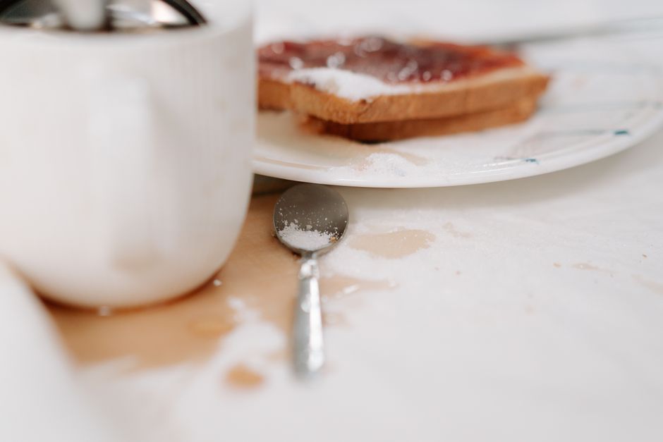 A close-up view of a kitchen countertop with flour scattered across its surface. Visible objects include a white ceramic mug on the left, a stainless steel measuring spoon lying flat with some flour on it, and a white plate in the background holding a piece of toast with jam. The scene is well-lit, highlighting the cleanliness of the countertop despite the flour mess, which may require surface cleaning. The setting appears to be a domestic kitchen space, emphasizing the importance of thorough cleaning and sanitisation, as offered by Greenwich Carpet Cleaning, in maintaining hygiene during everyday activities.