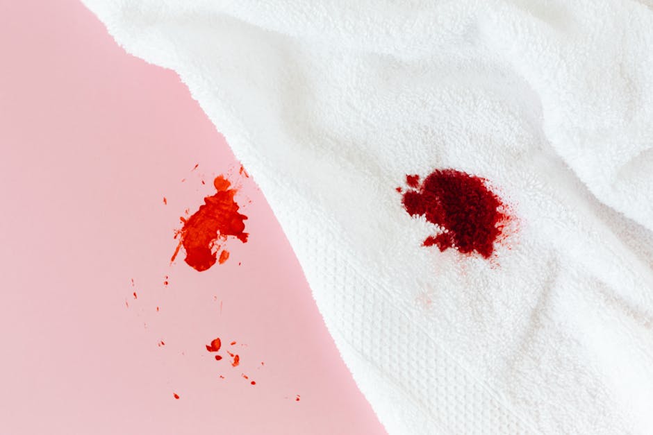 A white paper towel with a textured surface displaying a red stain near the center, indicating a fresh or dried spill. The background features a pink surface with a smaller red blotch nearby, suggesting accidental staining. The lighting is even, highlighting the contrast between the white paper and the vibrant color of the stain. This image exemplifies the need for effective surface cleaning and stain removal, relevant to domestic cleaning and sanitisation services offered by Greenwich Carpet Cleaning, as discussed on the page about emergency stain removal in Greenwich.